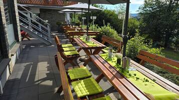 Un patio al aire libre con bancos y mesas de madera, algunas con cojines verdes, y sombrillas que dan sombra.