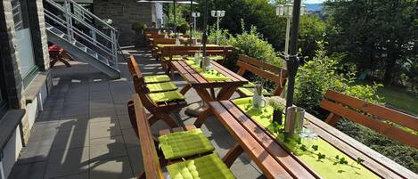 Un patio al aire libre con bancos y mesas de madera, algunas con cojines verdes, y sombrillas que dan sombra.