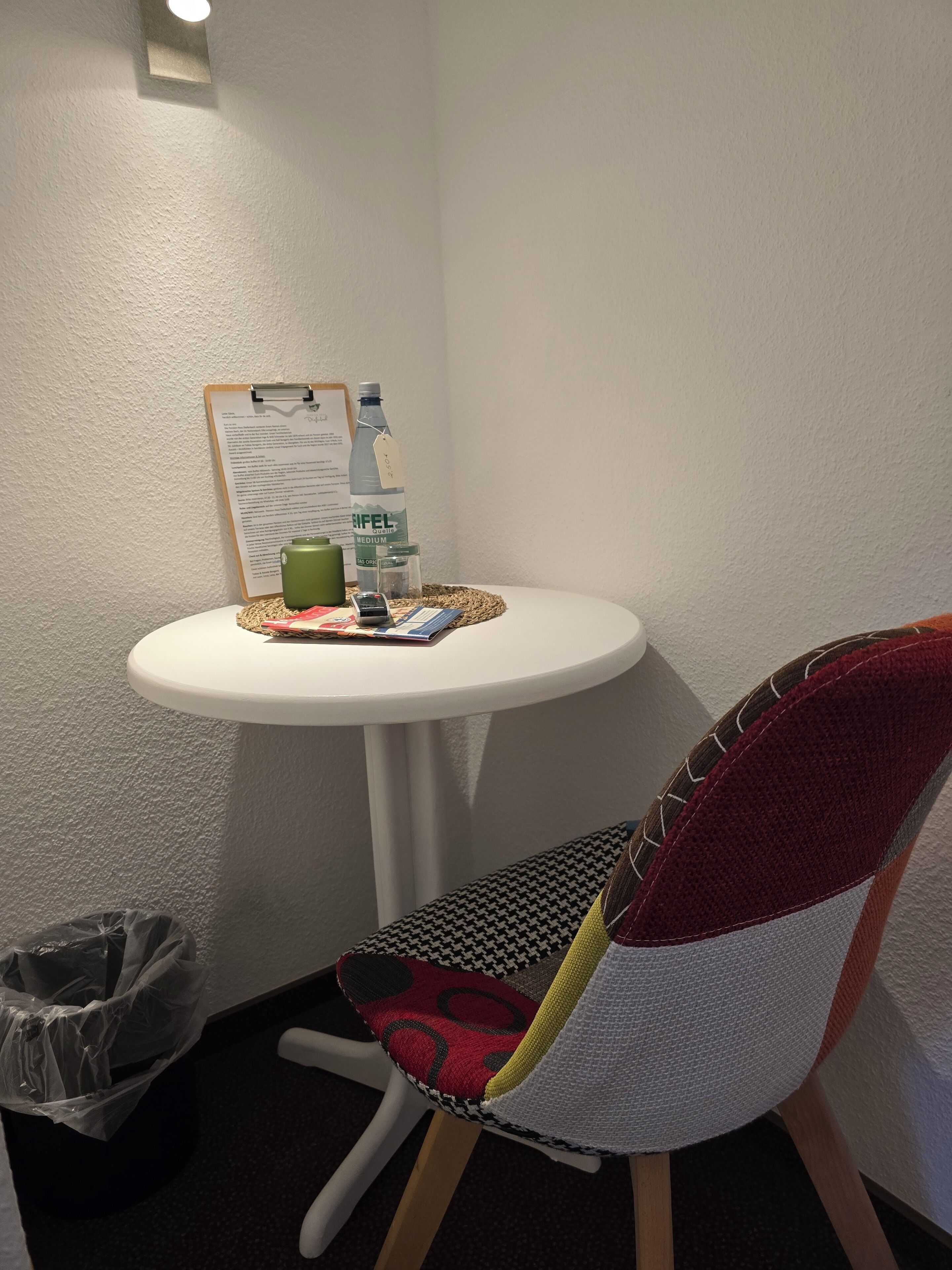 Une petite table blanche, sur laquelle se trouvent un presse-papiers, une bouteille d’eau, un thermos vert et un téléphone portable. Une chaise avec un coussin à motifs est placée devant la table.