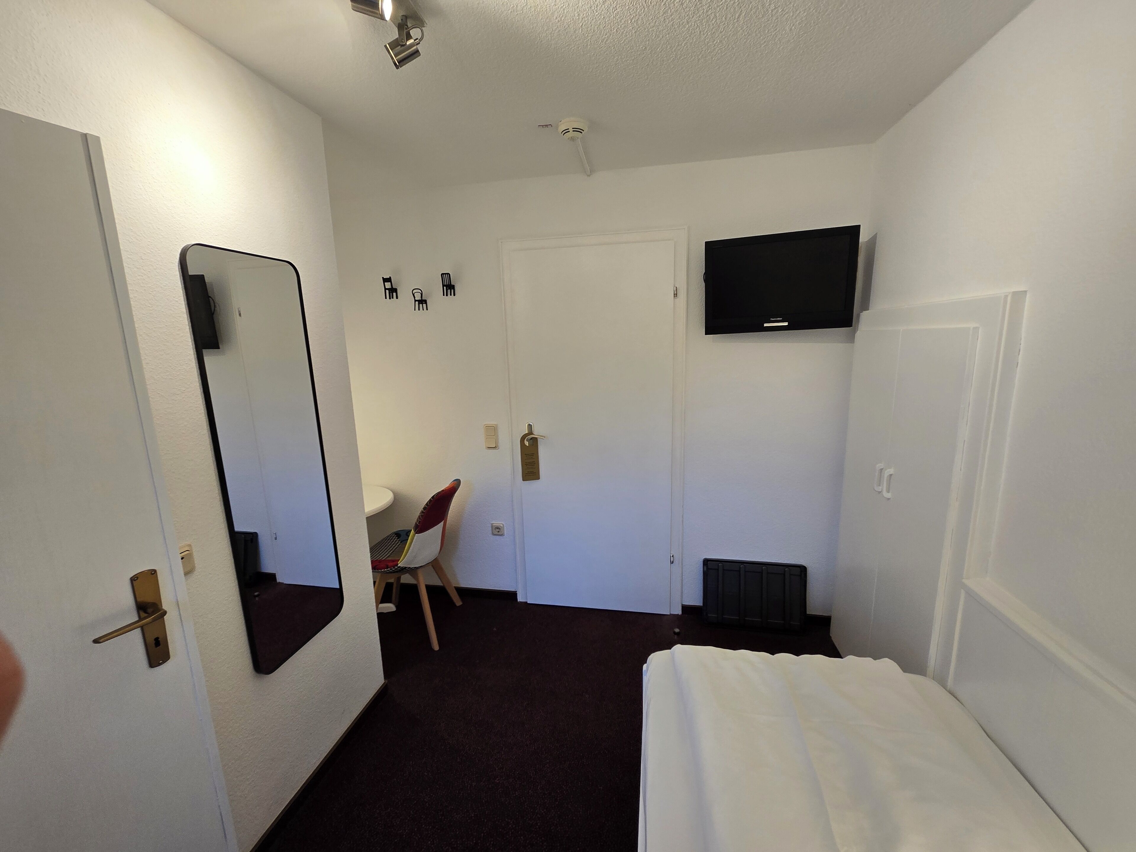 A small, well-lit room with a bed, a desk, a chair, a television mounted on the wall, and a door with a handle.