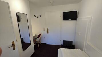 A small, well-lit room with a bed, a desk, a chair, a television mounted on the wall, and a door with a handle.