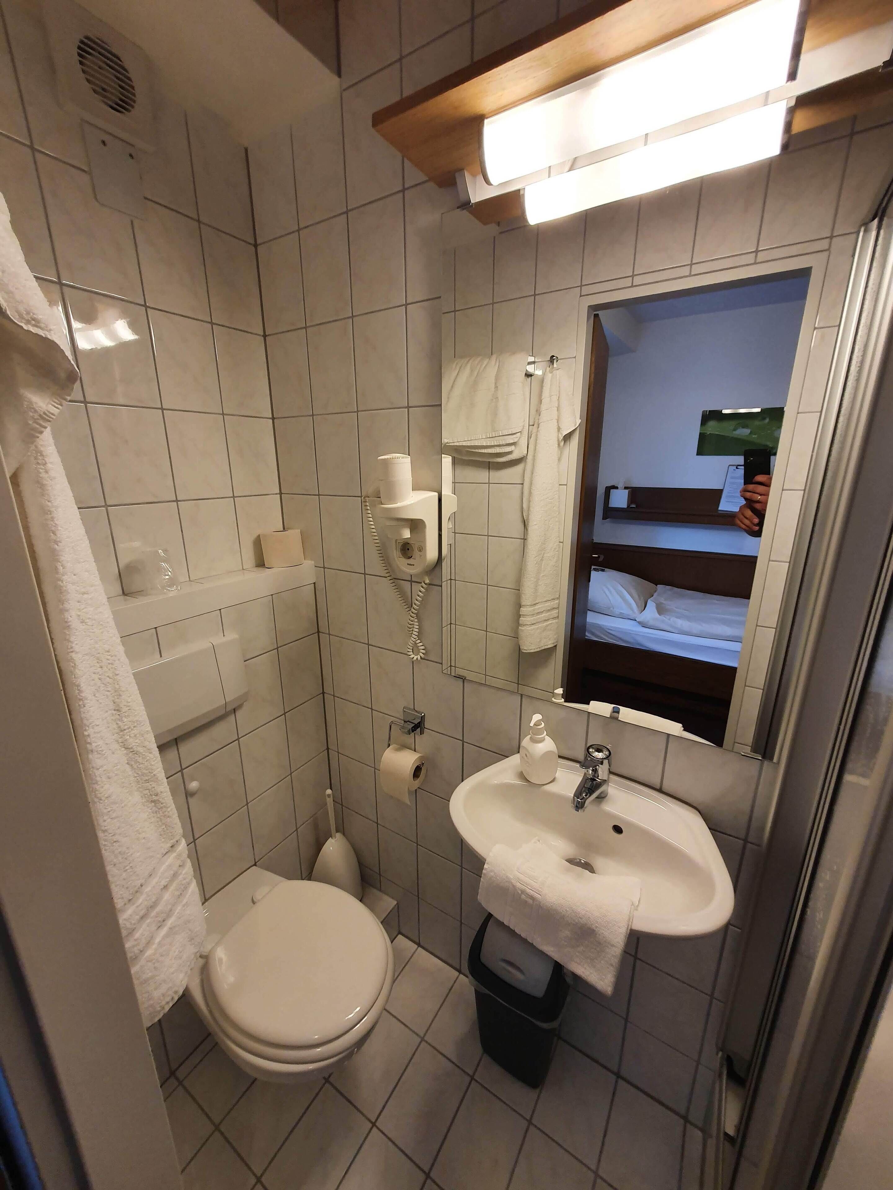 Baño pequeño y funcional con inodoro, lavamanos y ducha. Una toalla cuelga en la pared, y un espejo refleja una cama.