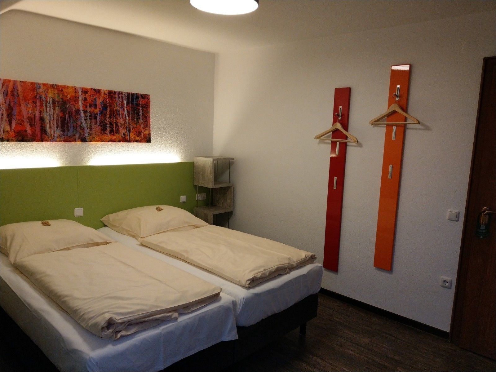 A double bed with a green headboard, a red and orange wall decoration, and a wooden hanger on the wall.