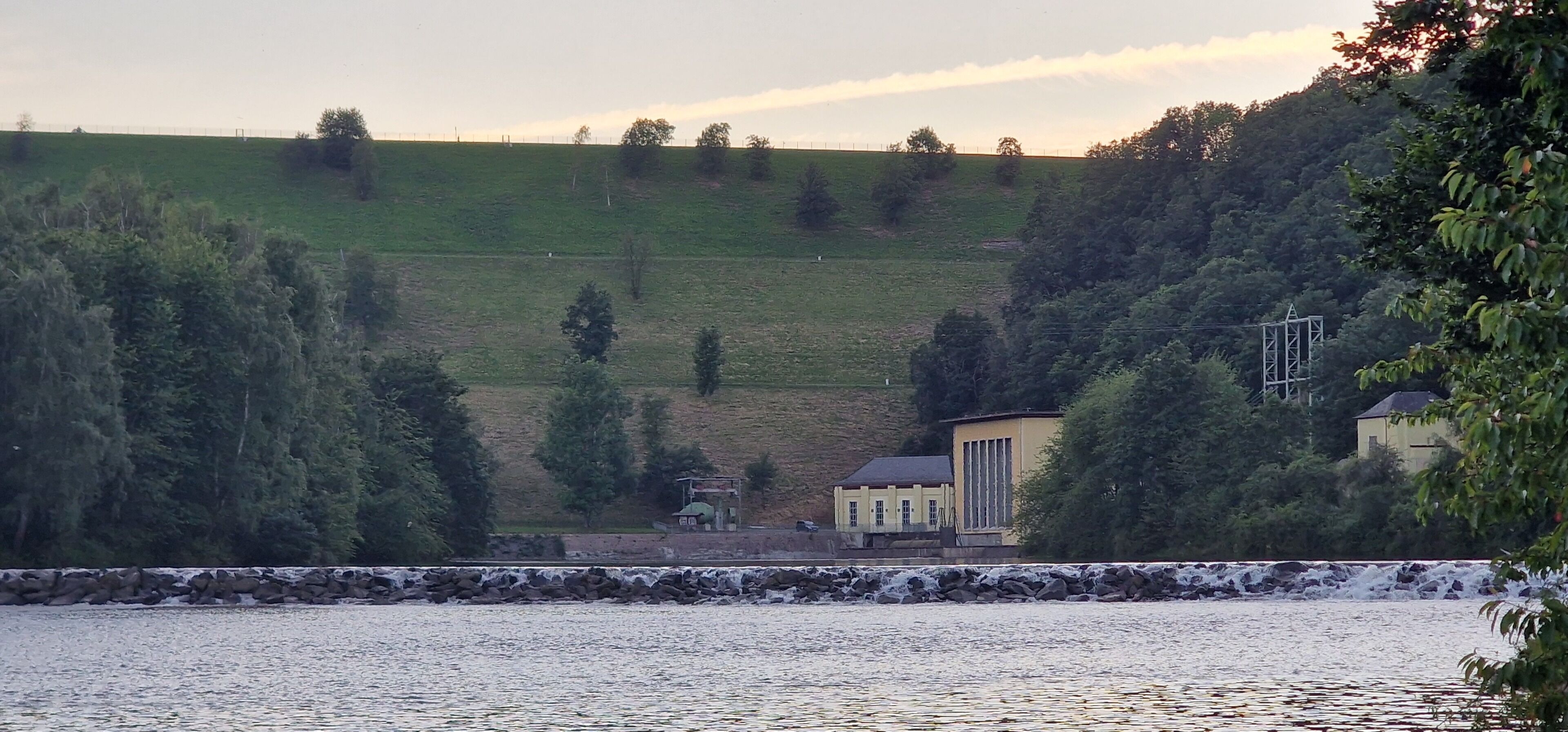 Une vue sur la rivière, avec un mur de pierre, un petit bâtiment et une colline parsemée d’arbres.