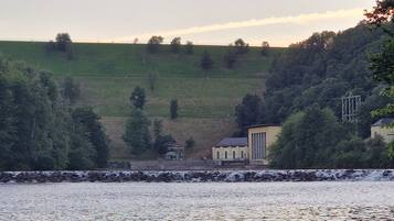 Une vue sur la rivière, avec un mur de pierre, un petit bâtiment et une colline parsemée d’arbres.