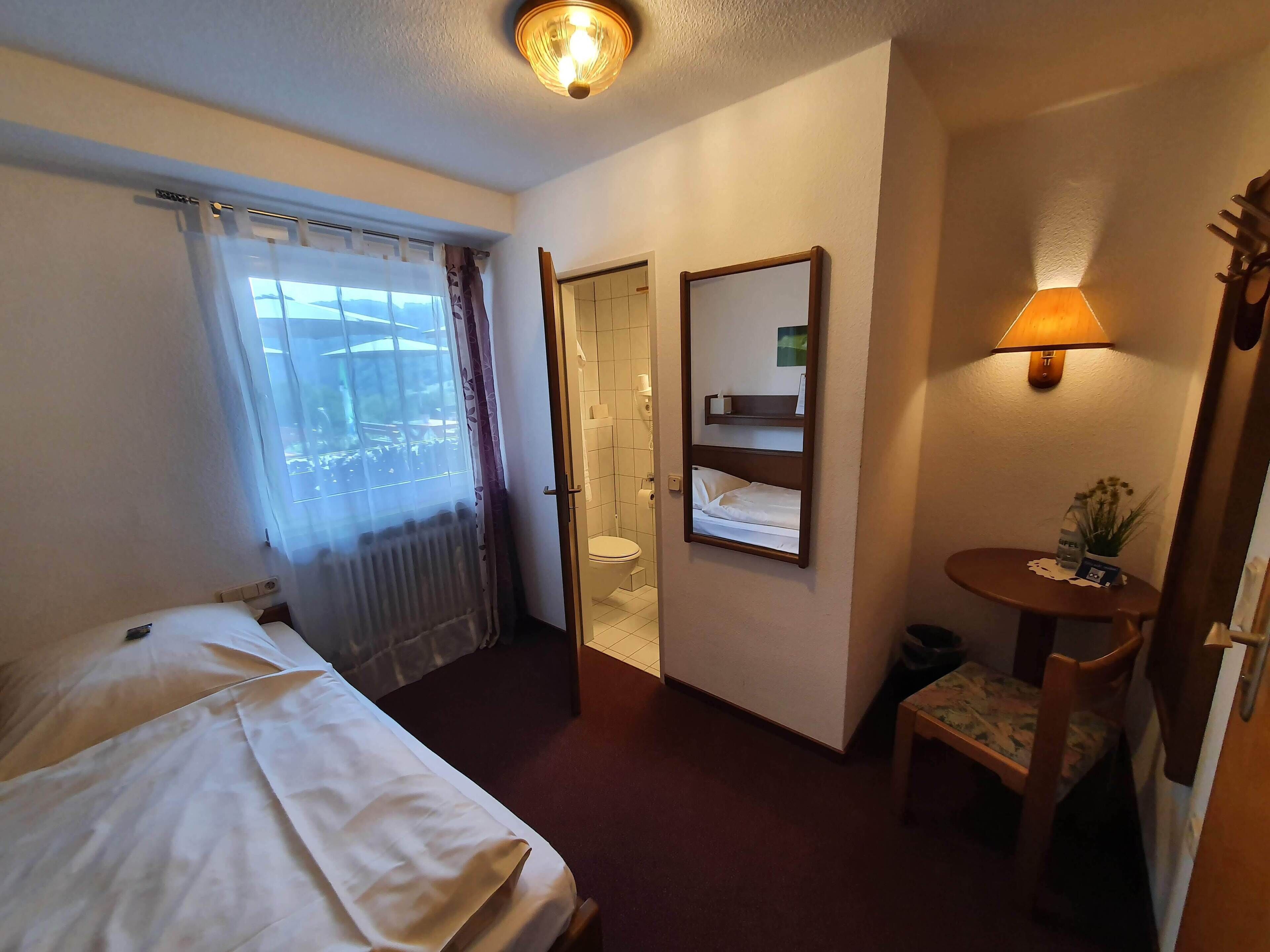 A hotel room with a bed, a small table with a lamp, a mirror, and a view of the outside through a window.