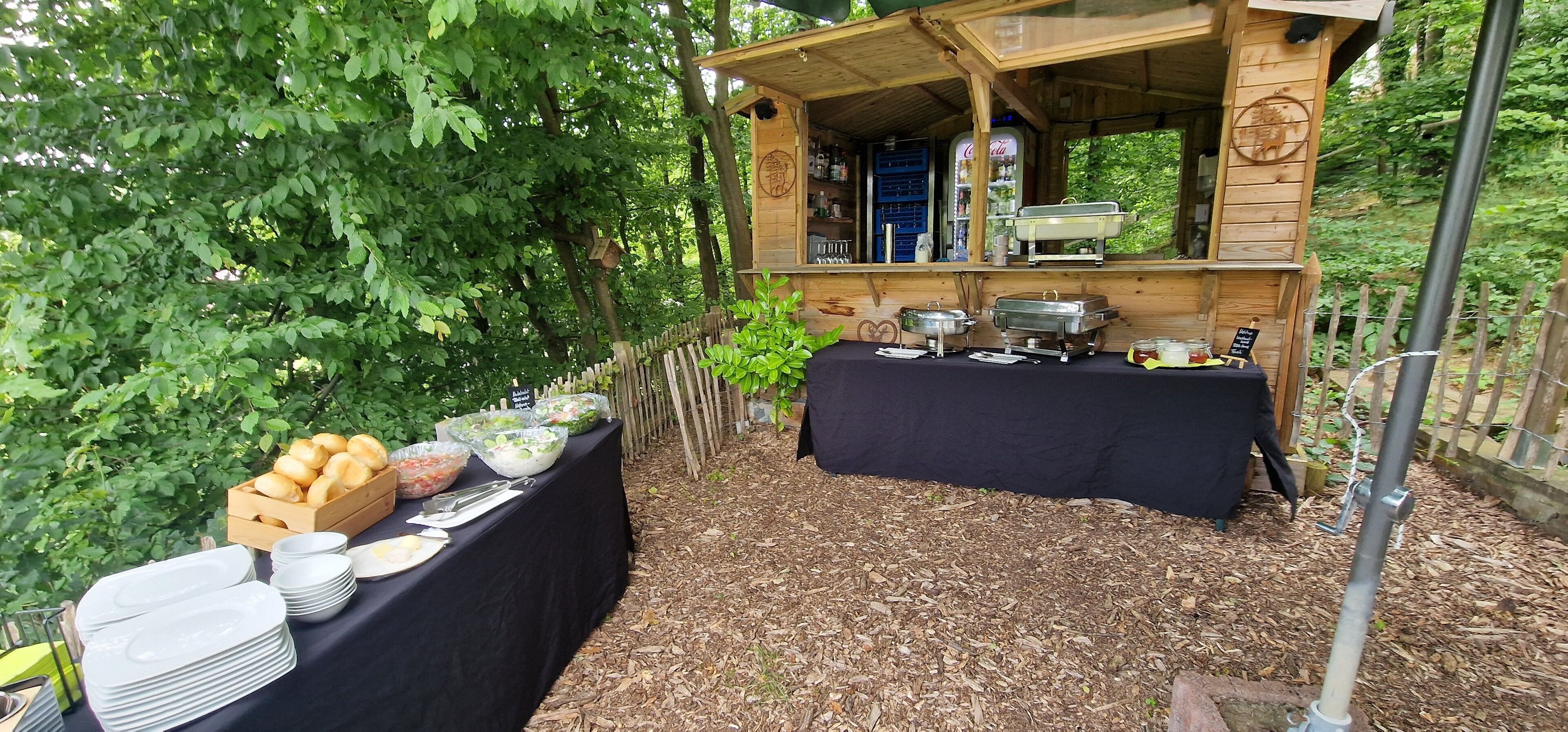 Un buffet en plein air, avec des tables sur lesquelles sont disposés des récipients contenant de la nourriture et des boissons, le tout entouré d’arbres.
