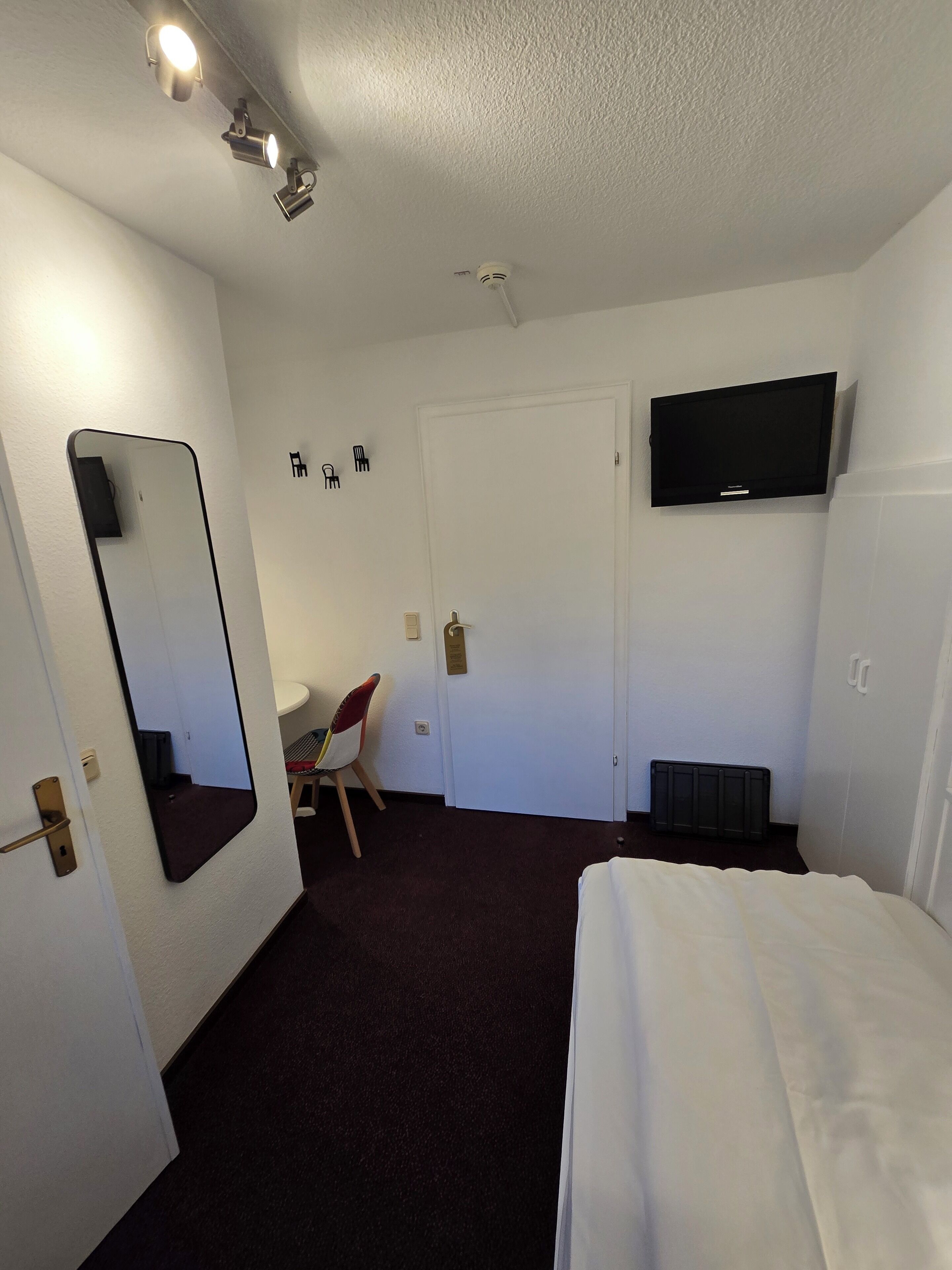 Habitación de hotel con cama, televisor montado en la pared, espejo, puerta y una silla.