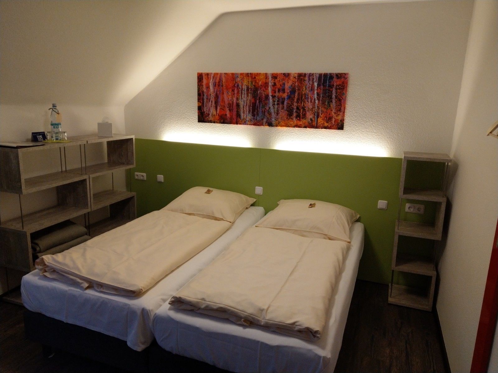 A room with two beds, a green headboard, a shelf with a bottle, and a painting on the wall.