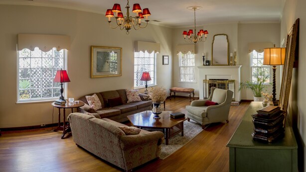 A spacious living room with a large sofa, a smaller armchair, a coffee table, and a fireplace. There are multiple windows providing natural light, and a chandelier hangs from the ceiling. - La Villa de Adelina (Ensenada)