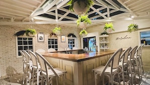 A bar/lounge area with a central wooden bar, white wicker chairs, hanging plants, and a brick wall with framed pictures. - La Villa de Adelina (Ensenada)