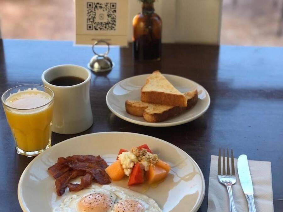 Een ontbijttafel met een bord eieren, spek en fruit, een glas sinaasappelsap, een kop koffie en een menubord.