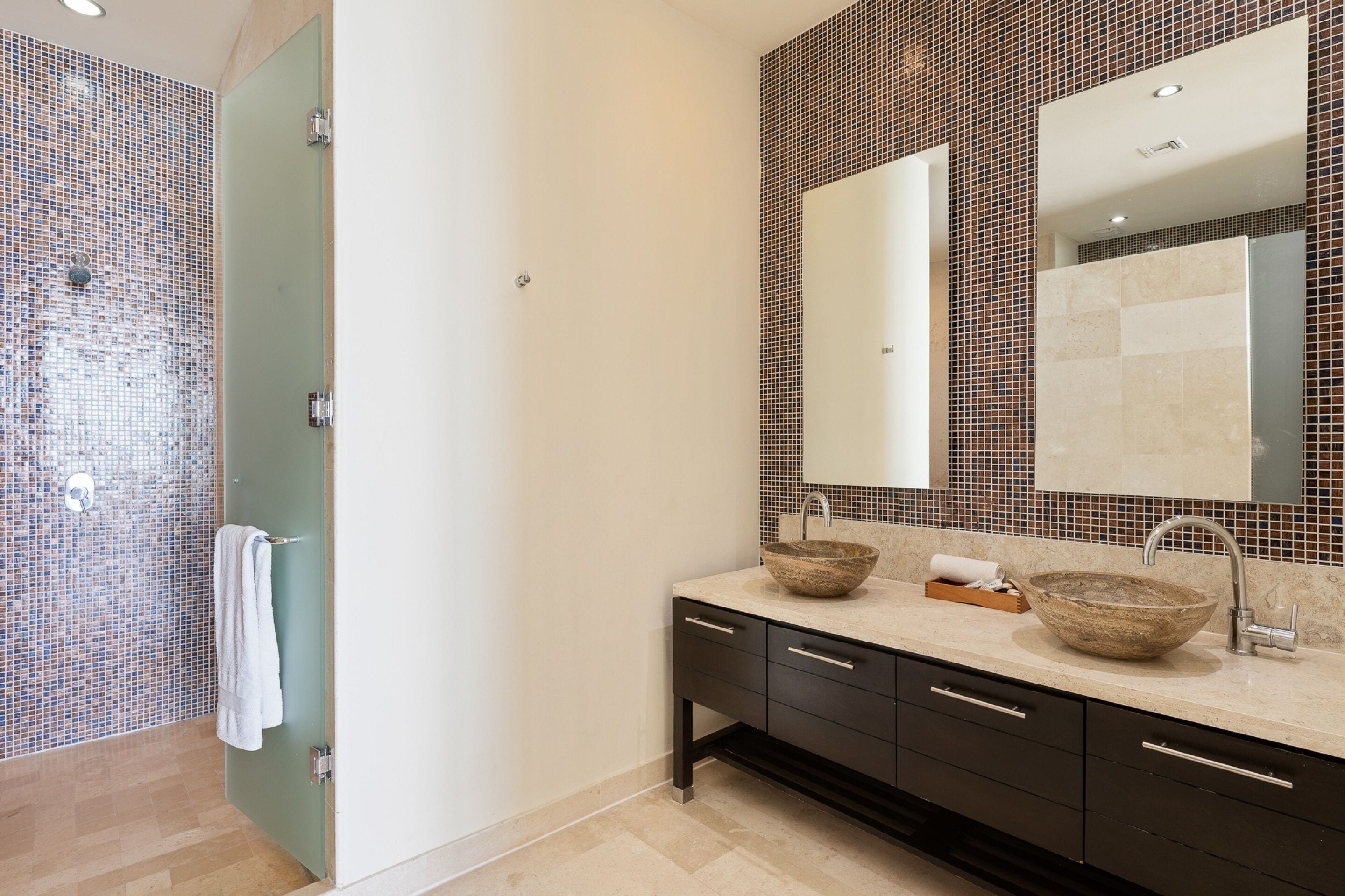 A modern bathroom with a mosaic tile shower wall, a large mirror, and a wooden bowl sink.