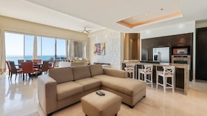 A modern living room with a large sectional sofa, a dining area with a table and chairs, and a kitchen with dark cabinetry and a large island. - Encanto Living 1201 -4BR by Kivoya (Puerto Peñasco)