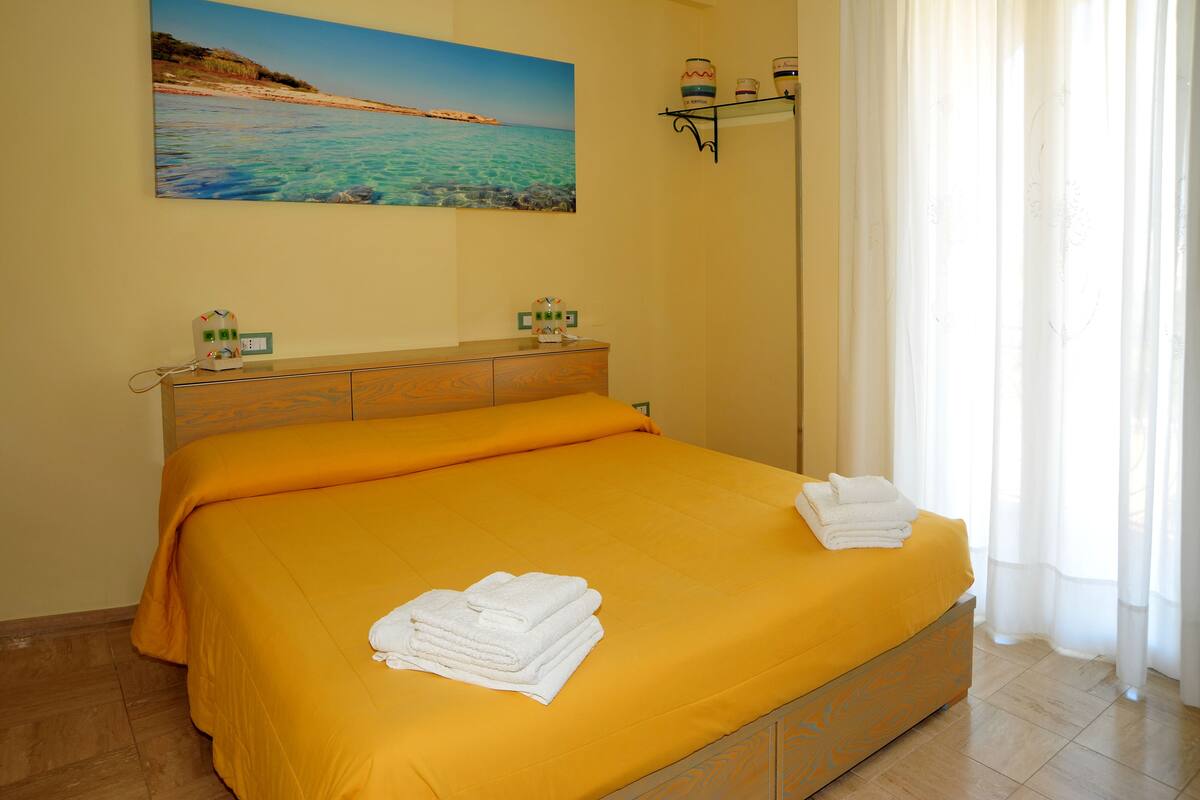 A hotel room with a bed, a nightstand, a painting of a beach, and a window with curtains.