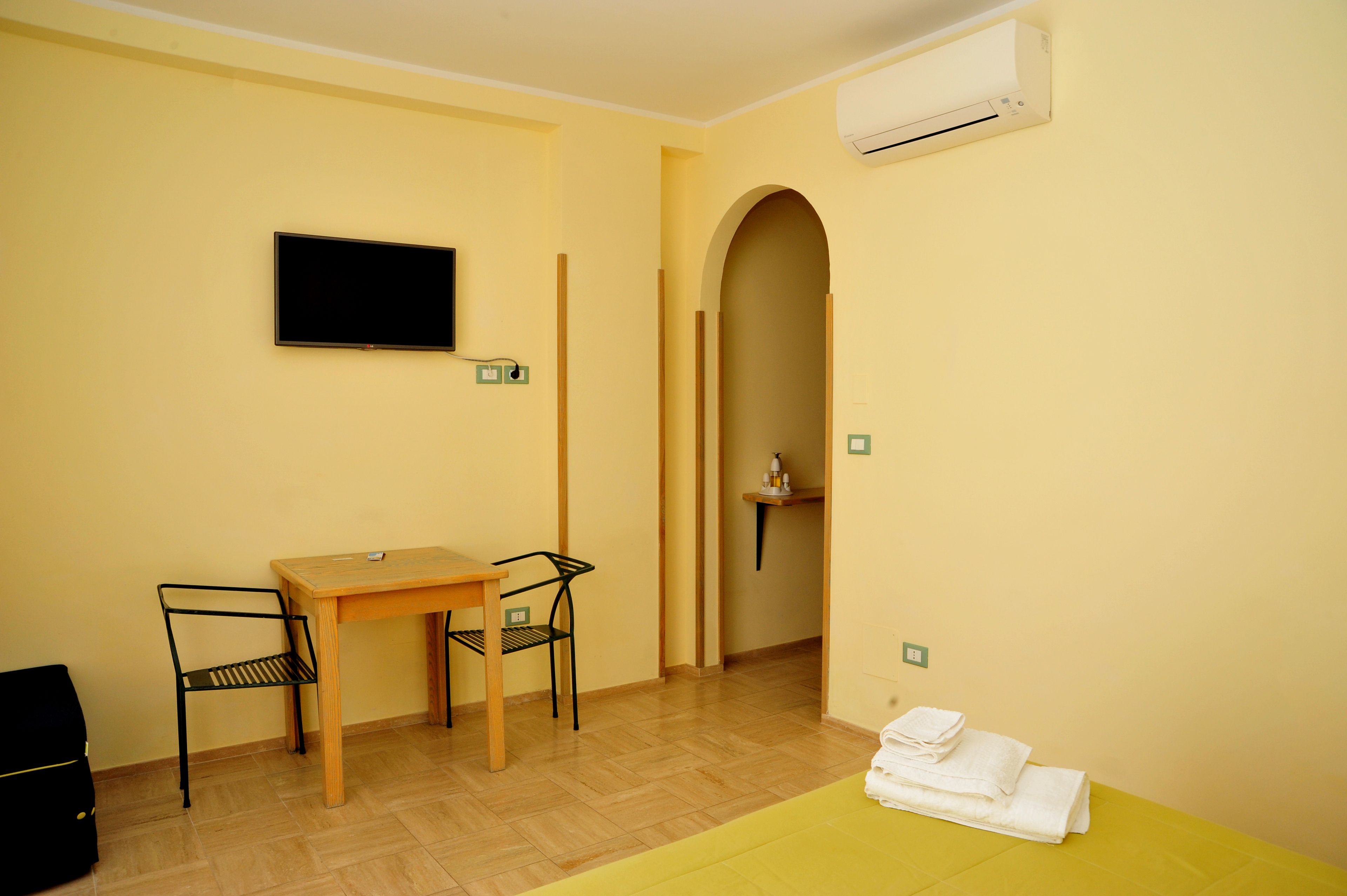 A room with a wooden table, two chairs, a TV mounted on the wall, an air conditioning unit, and a towel on the floor.