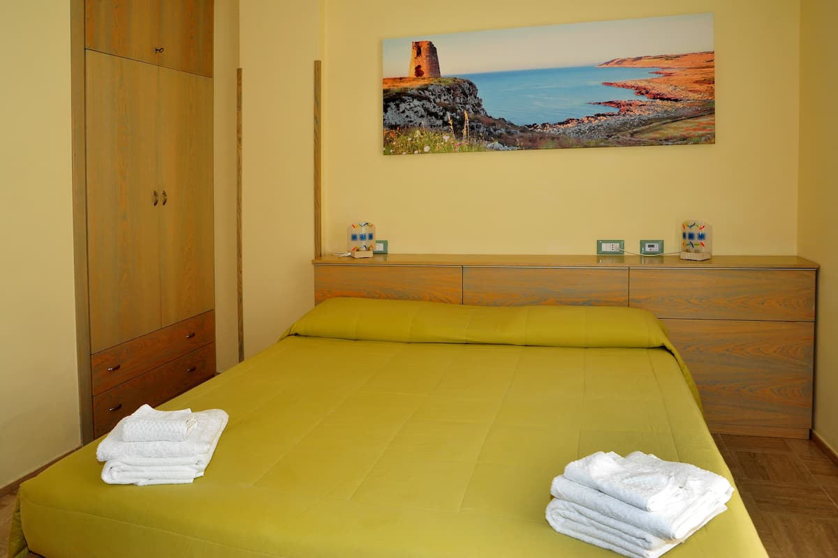 A hotel room with a bed, a wooden wardrobe, a painting of a coastal landscape, and a small table with a clock and a decorative item.
