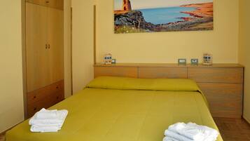 A hotel room with a bed, a wooden wardrobe, a painting of a coastal landscape, and a small table with a clock and a decorative item.