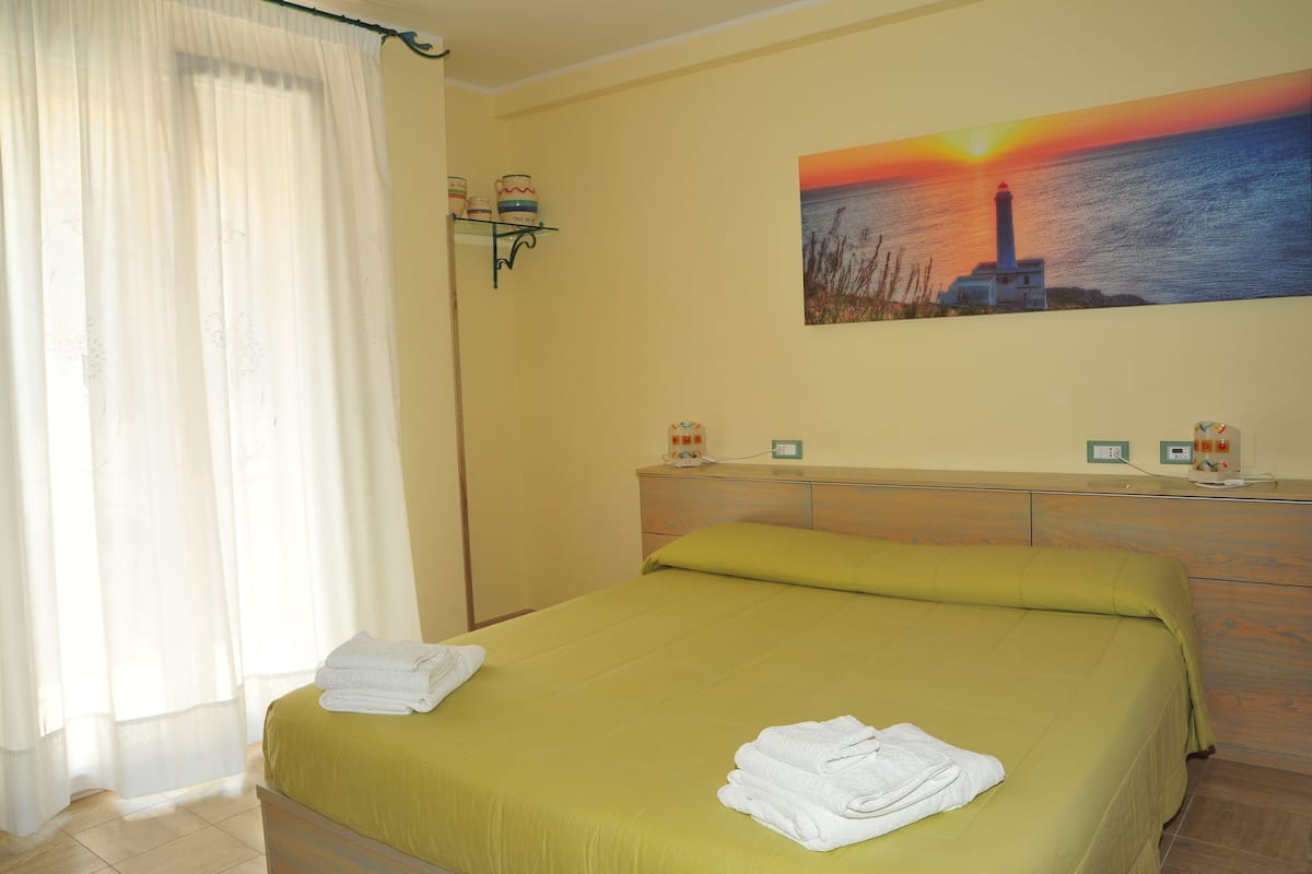 A neatly made bed with a green bedspread and white towels, a painting of a lighthouse on the wall, and a bedside table with a lamp and a clock.