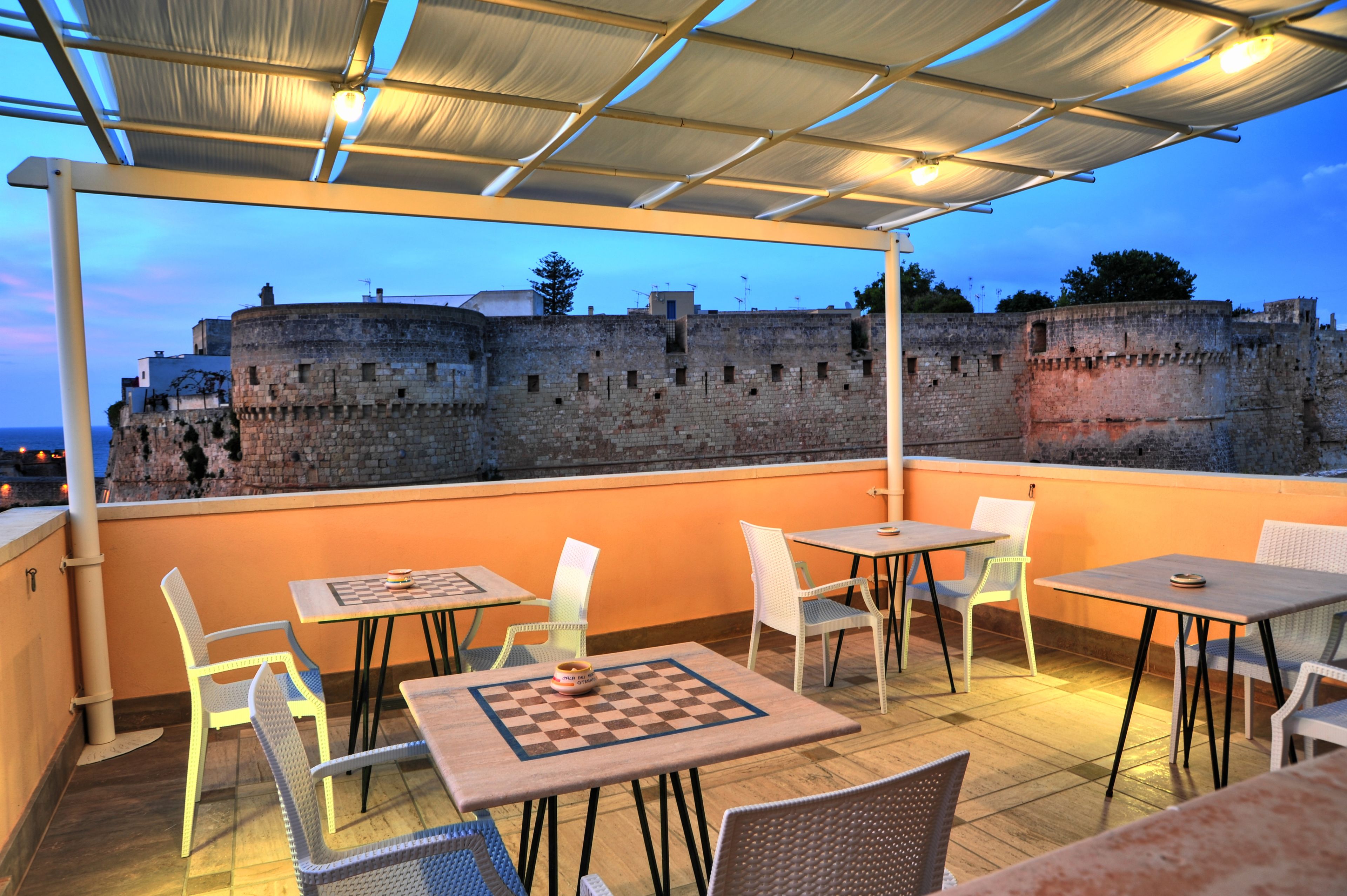 Terraço ao ar livre com mesas, cadeiras, um tabuleiro de xadrez e vista para uma parede de pedra histórica.