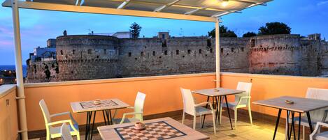 Terraço ao ar livre com mesas, cadeiras, um tabuleiro de xadrez e vista para uma parede de pedra histórica.