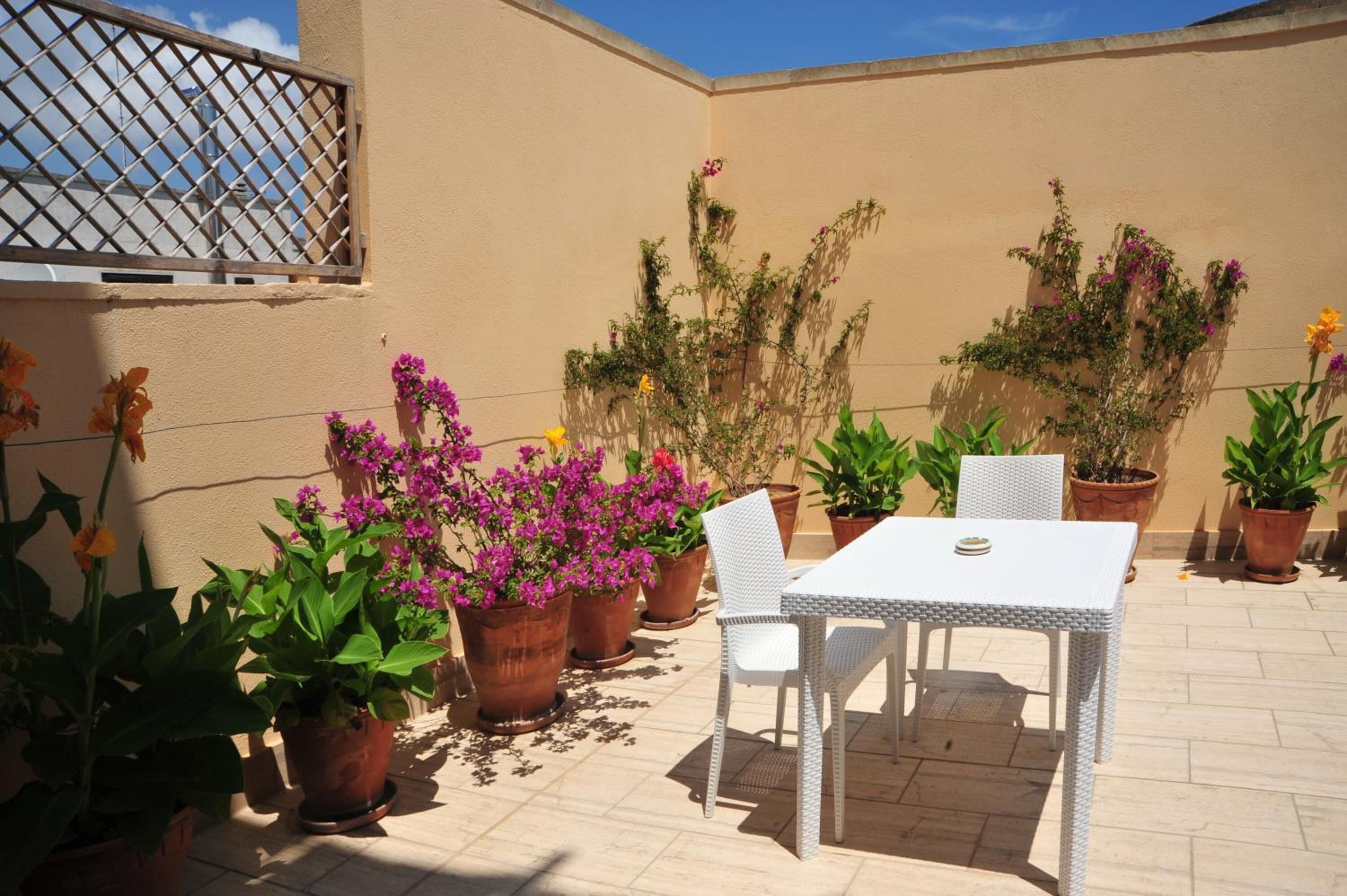 Un patio al aire libre con una mesa y sillas blancas, rodeado de macetas con plantas y flores.