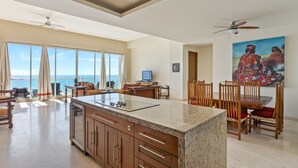 A spacious kitchen with a large island, dining area with wooden chairs, and a painting on the wall. - Encanto Living 1202 -4BR by Kivoya (Puerto Peñasco)