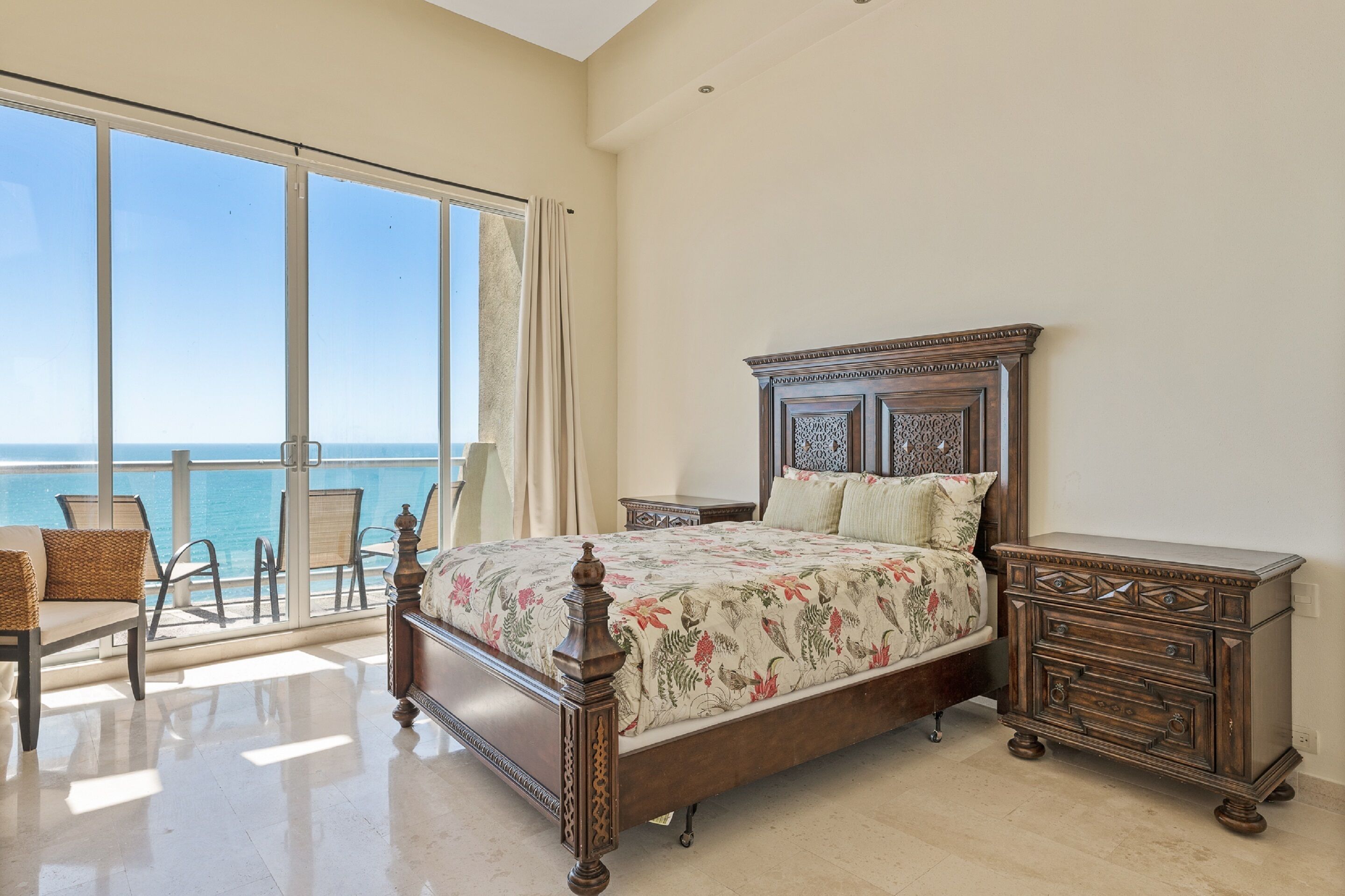 Ein Schlafzimmer mit großem Fenster zum Meer, einem hölzernen Bett mit Blumenmuster-Bettwäsche und einem Nachttisch mit Schubladen.