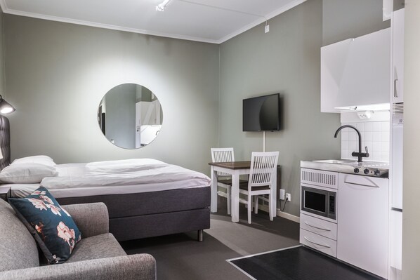 A compact hotel room with a bed, a small desk, a chair, a TV, a kitchenette, and a mirror.