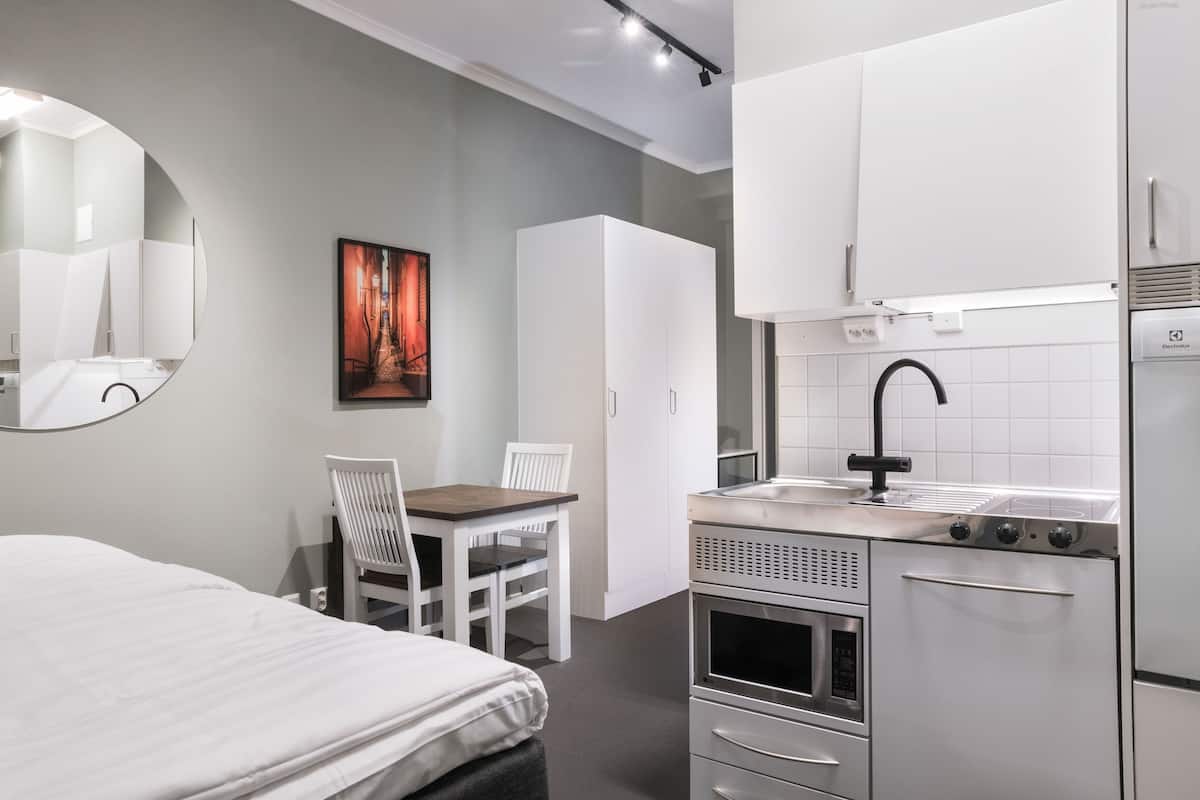 A modern hotel room with a bed, a dining area with a table and chairs, a kitchen with a sink and stove, and a large mirror on the wall.