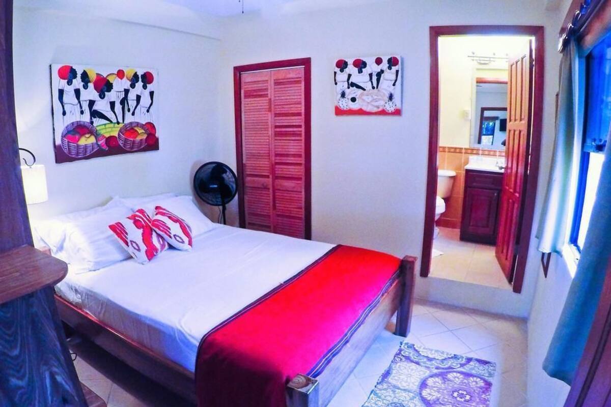 A bedroom with a bed, a red bedspread, a fan, and artwork on the walls.