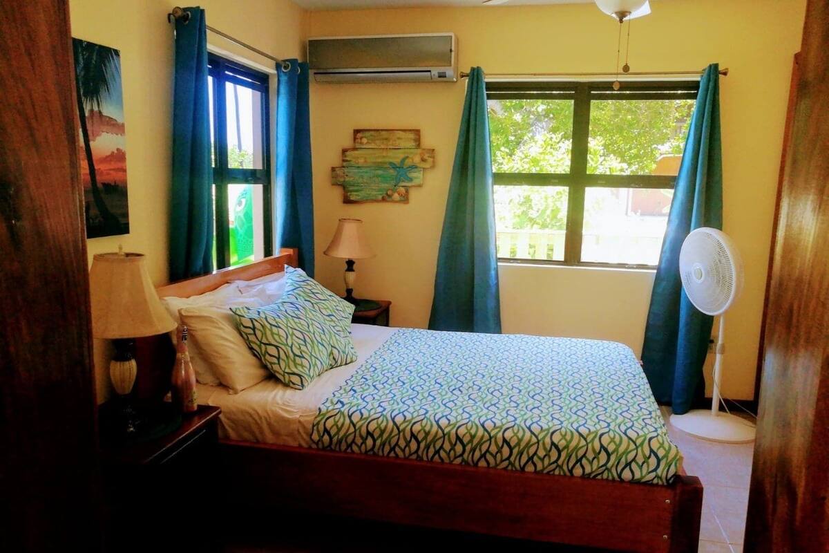 A bedroom with a bed, a nightstand, a lamp, a fan, and a window with curtains.