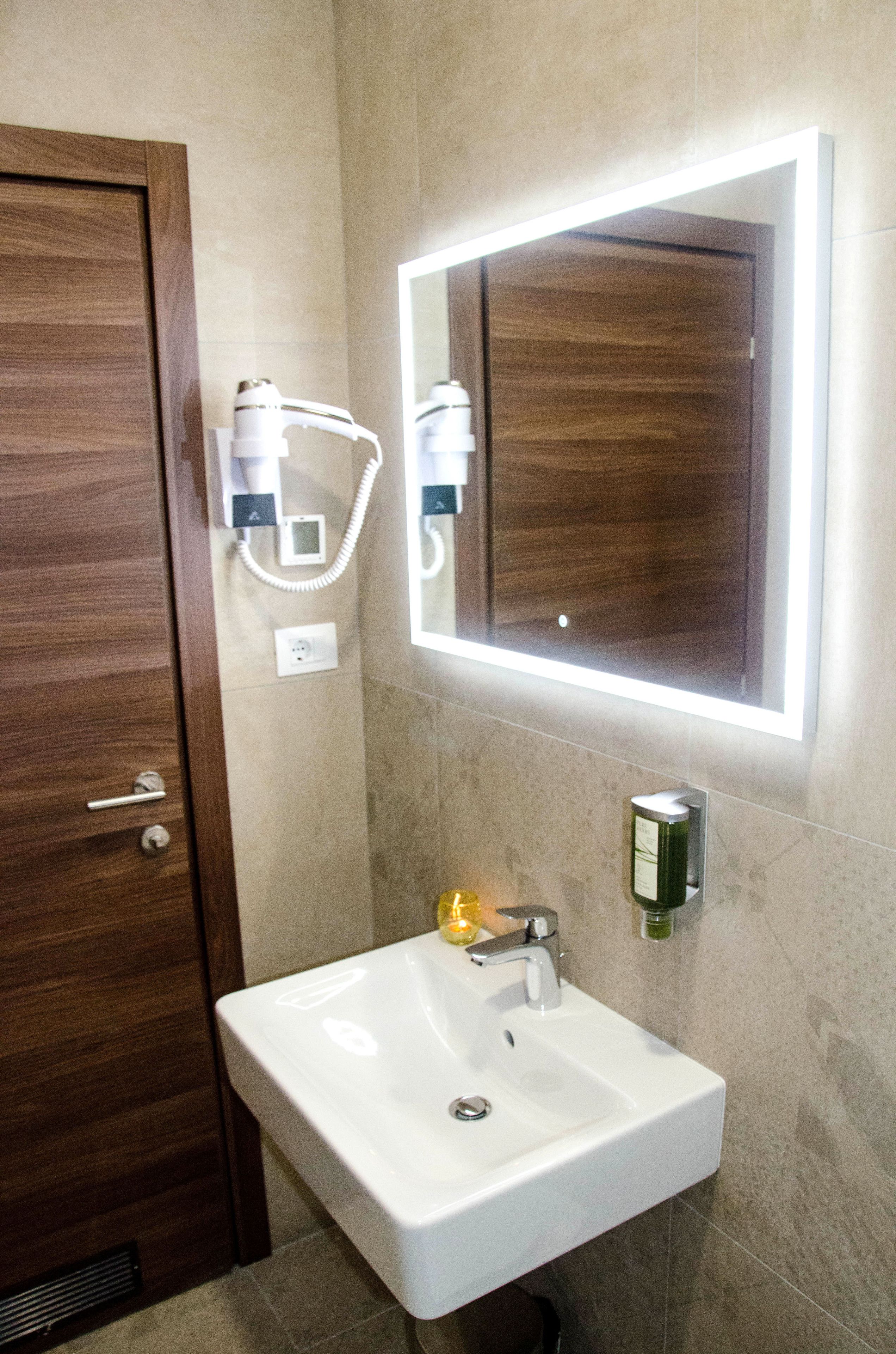 Ein Badezimmer mit Holztür, einem weißen Waschbecken, einem Spiegel mit integrierter Beleuchtung und einem an der Wand befestigten Föhn.