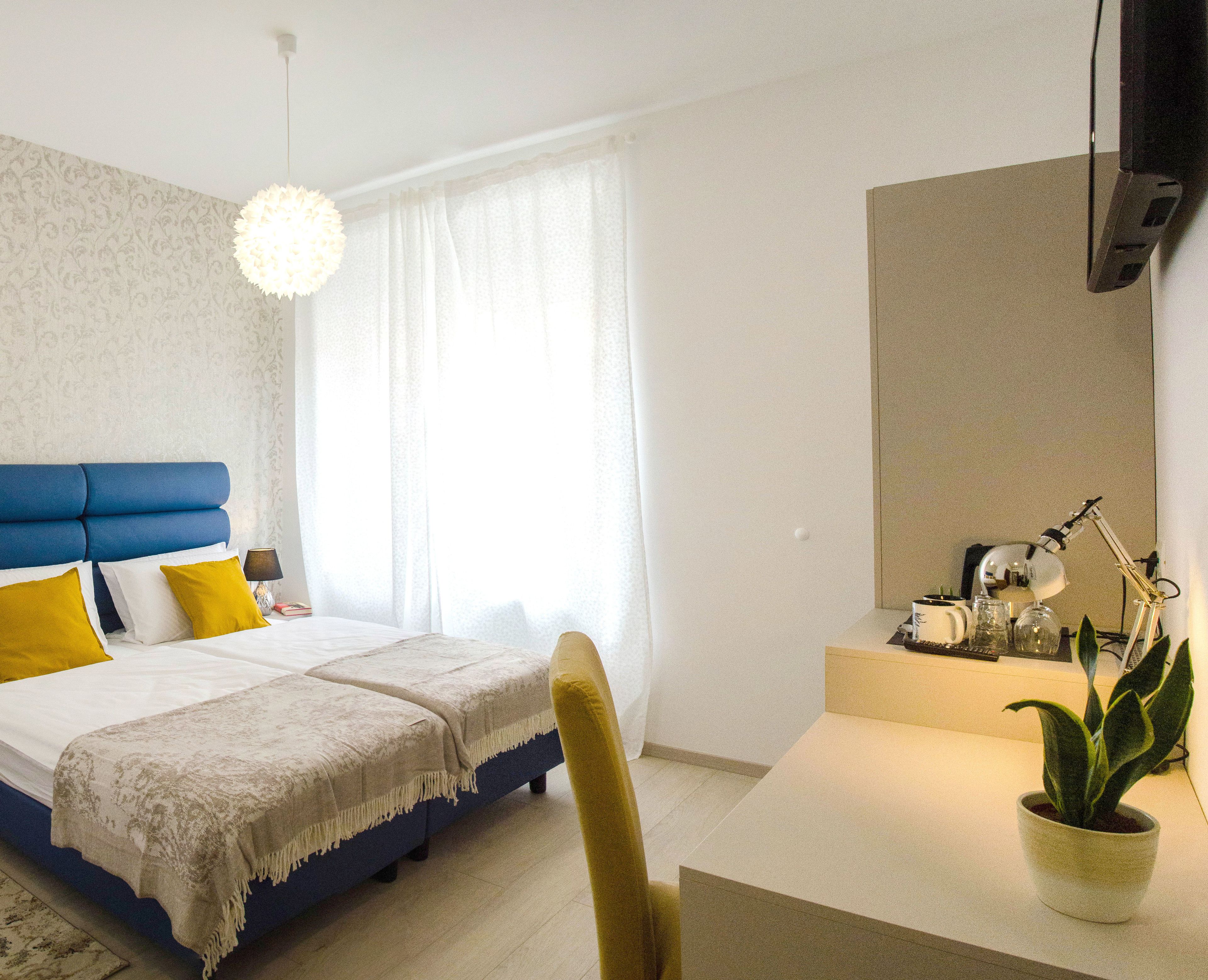 A modern hotel room with a blue headboard, a bed with white and yellow bedding, a desk with a potted plant, and a large window with sheer curtains.