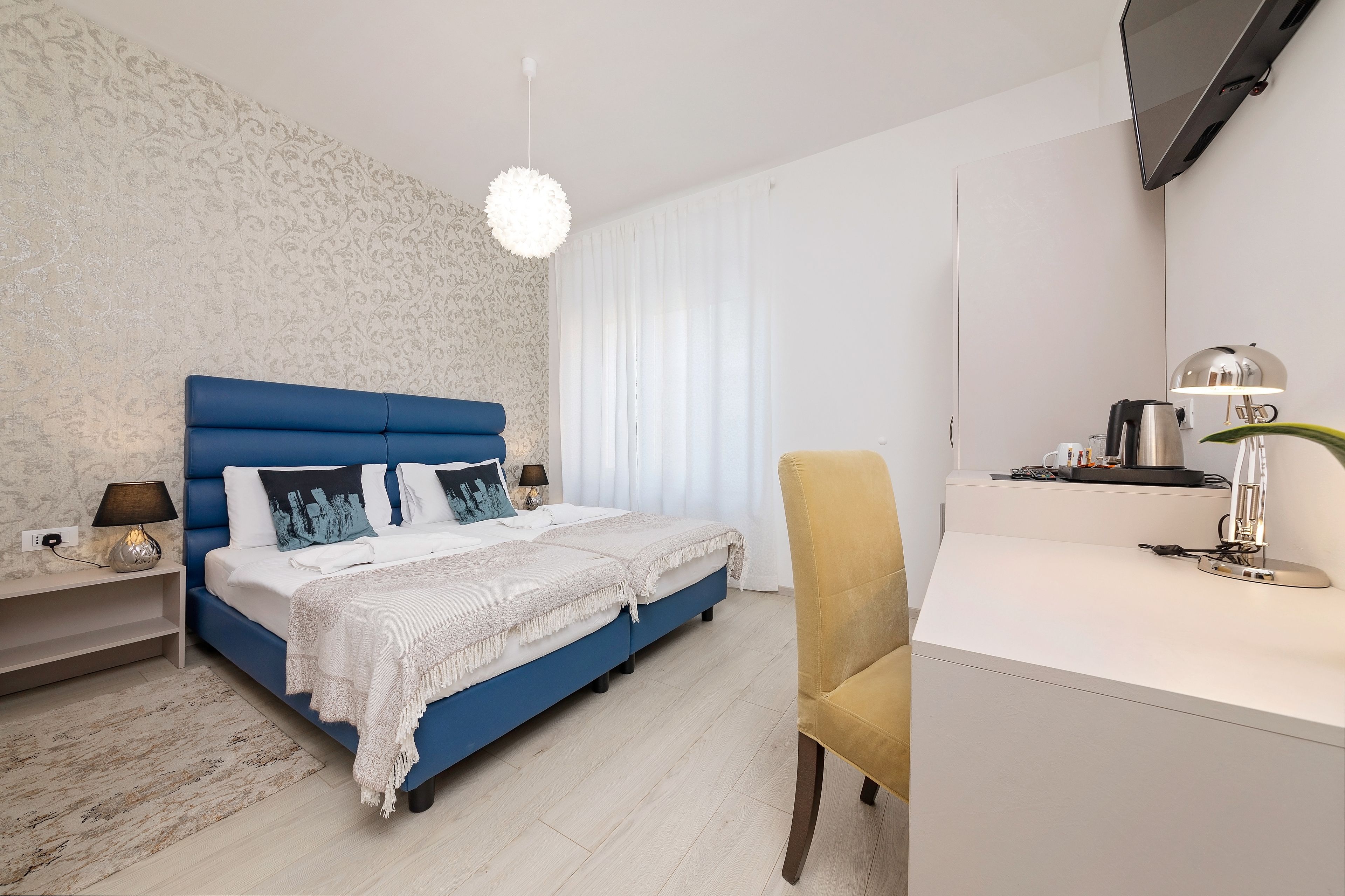 Un dormitorio moderno con una cama azul, ropa de cama blanca, un escritorio con lámpara y un televisor en la pared.