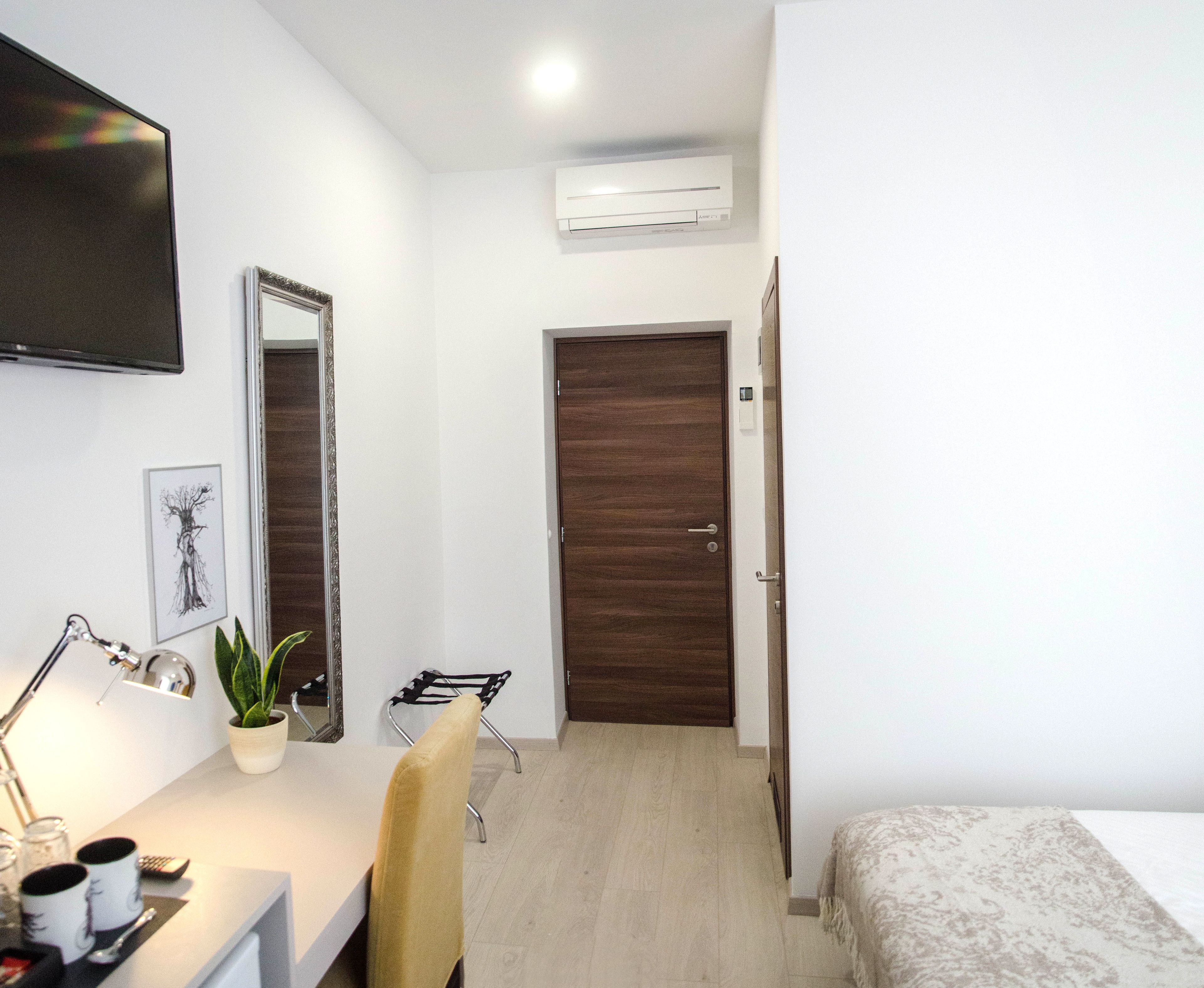 Une chambre d’hôtel avec un lit, un bureau avec une lampe, une chaise, une plante en pot, un téléviseur fixé au mur et une porte.