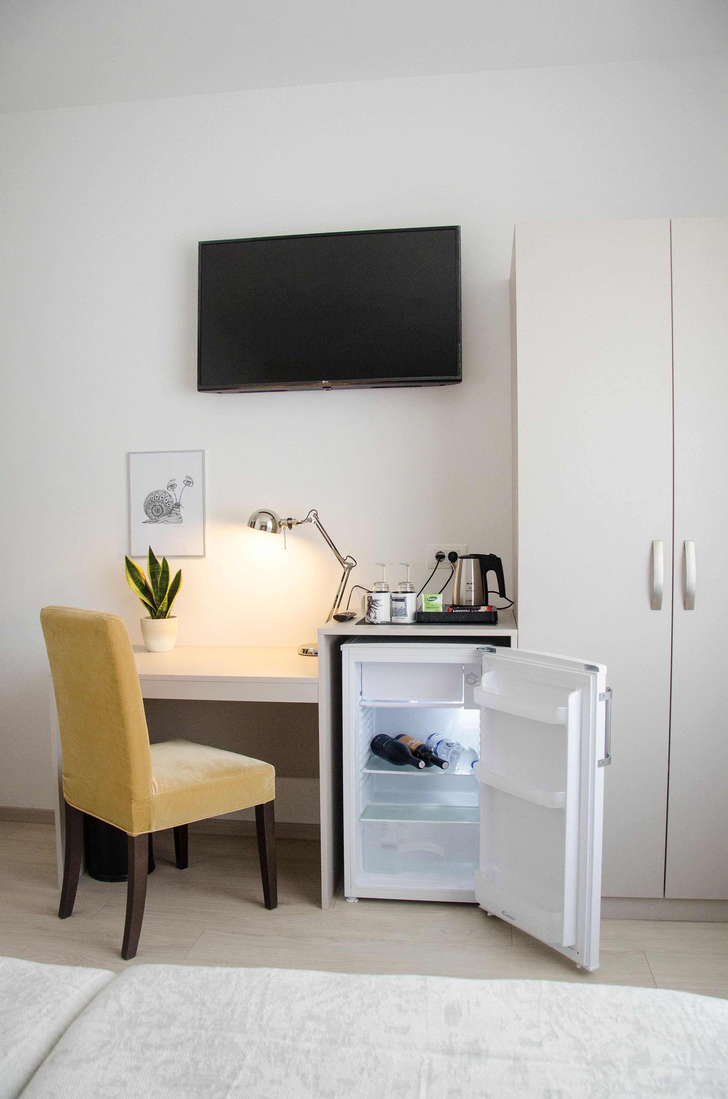 Une pièce de taille modeste, comprenant un bureau, une chaise, un téléviseur fixé au mur, un petit réfrigérateur et une plante en pot.