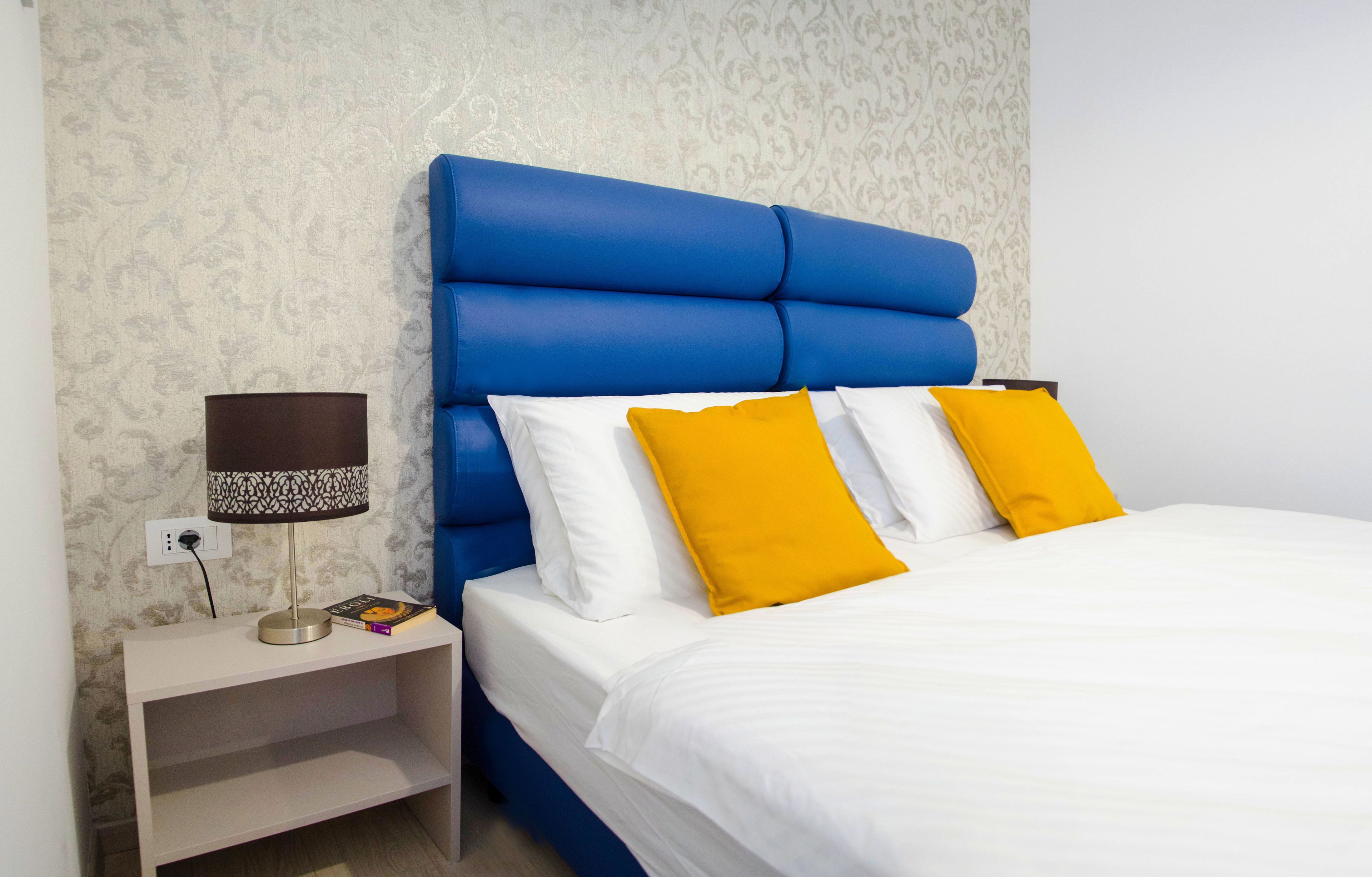 A neatly made bed with white and yellow pillows, a blue headboard, and a bedside table with a lamp and book.