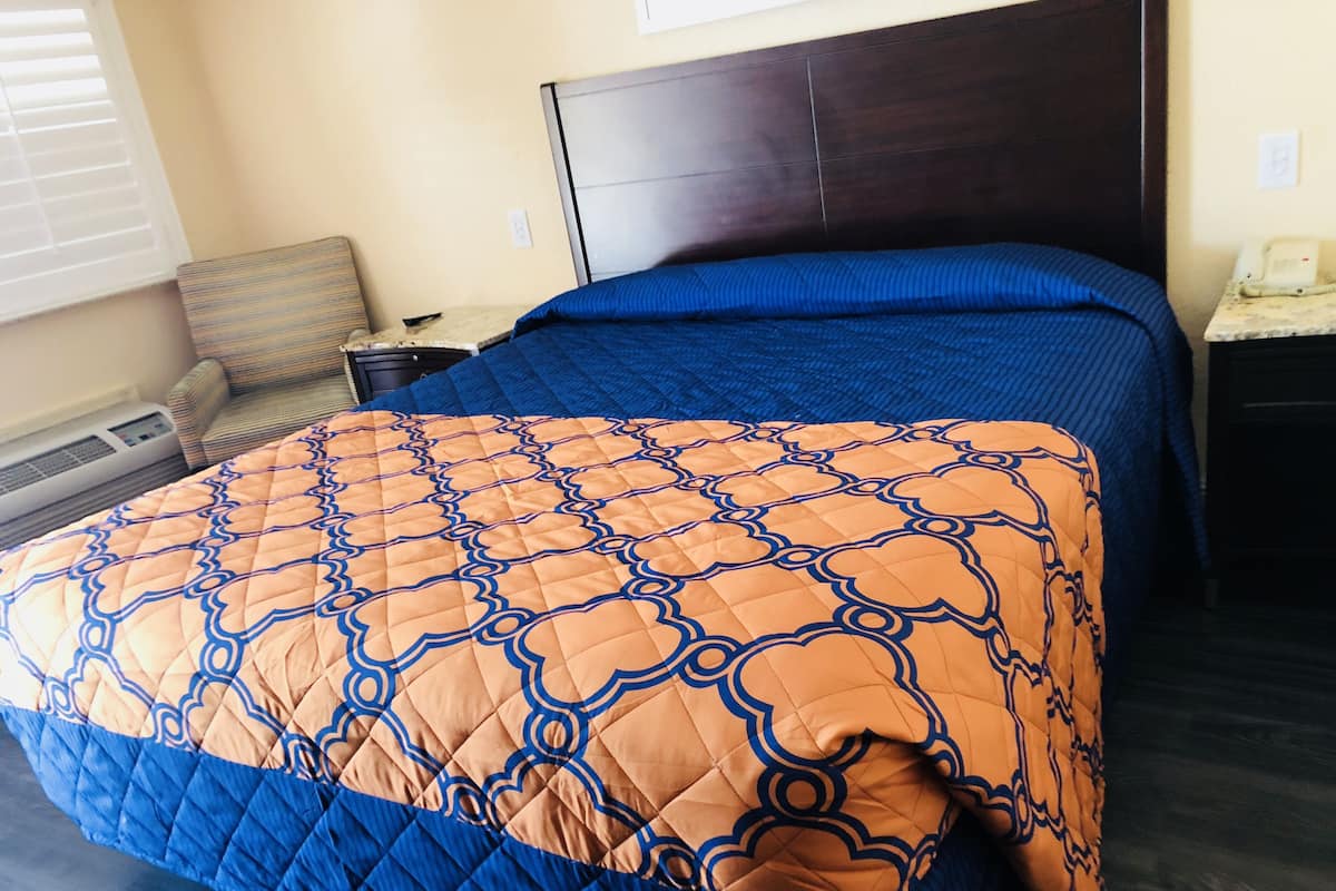 A bedroom with a dark wood headboard, a blue and orange quilted bedspread, a bedside table with a lamp, and a chair with a striped cushion.