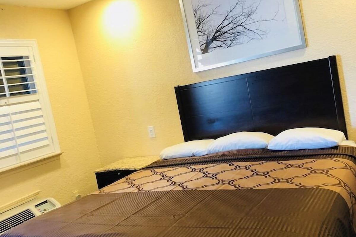 A bedroom with a bed, a headboard, a window with blinds, and a framed picture on the wall.