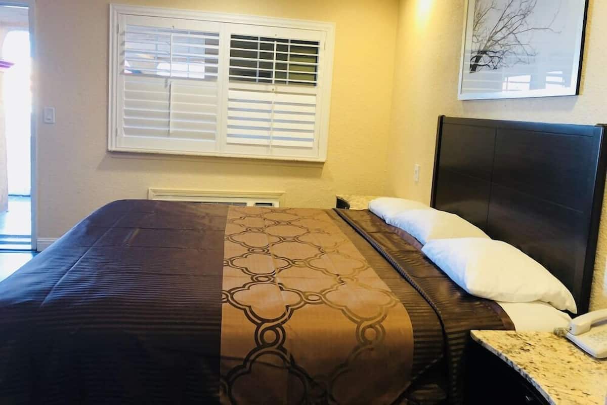 A hotel room with a bed, a headboard, a window with blinds, a wall-mounted phone, and a framed picture.