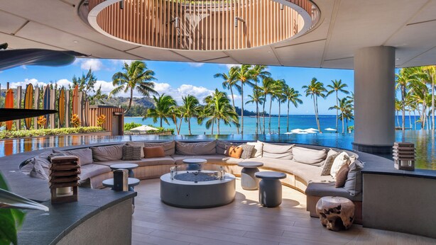 A modern lounge area with a curved seating arrangement, a central fire pit, and a view of the ocean and palm trees. - SUMMER REDUCED! Villa 102-103 Stunning Ocean Views at Turtle Bay (Kahuku)