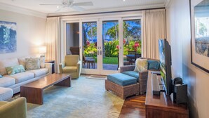 A spacious living room with a sofa, armchairs, a coffee table, and a TV. There is a sliding glass door leading to a balcony with outdoor furniture and plants. - SUMMER REDUCED! Villa 105-106 Ocean View Turtle Bay (Kahuku)