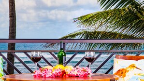 Uma mesa de madeira com uma garrafa de vinho, duas taças de vinho e um arranjo floral numa varanda com vista para o oceano.