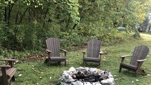 Un foyer entouré de chaises Adirondack et d’une table de pique-nique, le tout installé dans un espace herbeux bordé d’arbres.