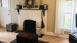 Un salon avec une cheminée en briques blanches, une étagère ornée d’objets décoratifs, un banc noir, une malle en bois et une fenêtre avec des rideaux.