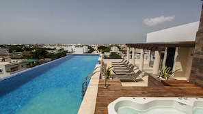 A rooftop infinity pool with a wooden deck and lounge chairs, a hot tub, and a view of the cityscape.