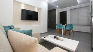 Een moderne woonkamer met een flatscreen televisie aan de muur, een bank met turquoise kussens, een salontafel met een decoratief object, en een eetgedeelte met turquoise stoelen.