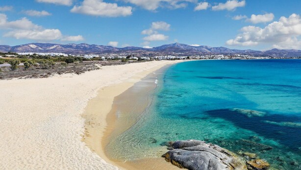 Une plage à l’eau turquoise et limpide, bordée d’un rivage sablonneux, avec une chaîne de montagnes en arrière-plan.