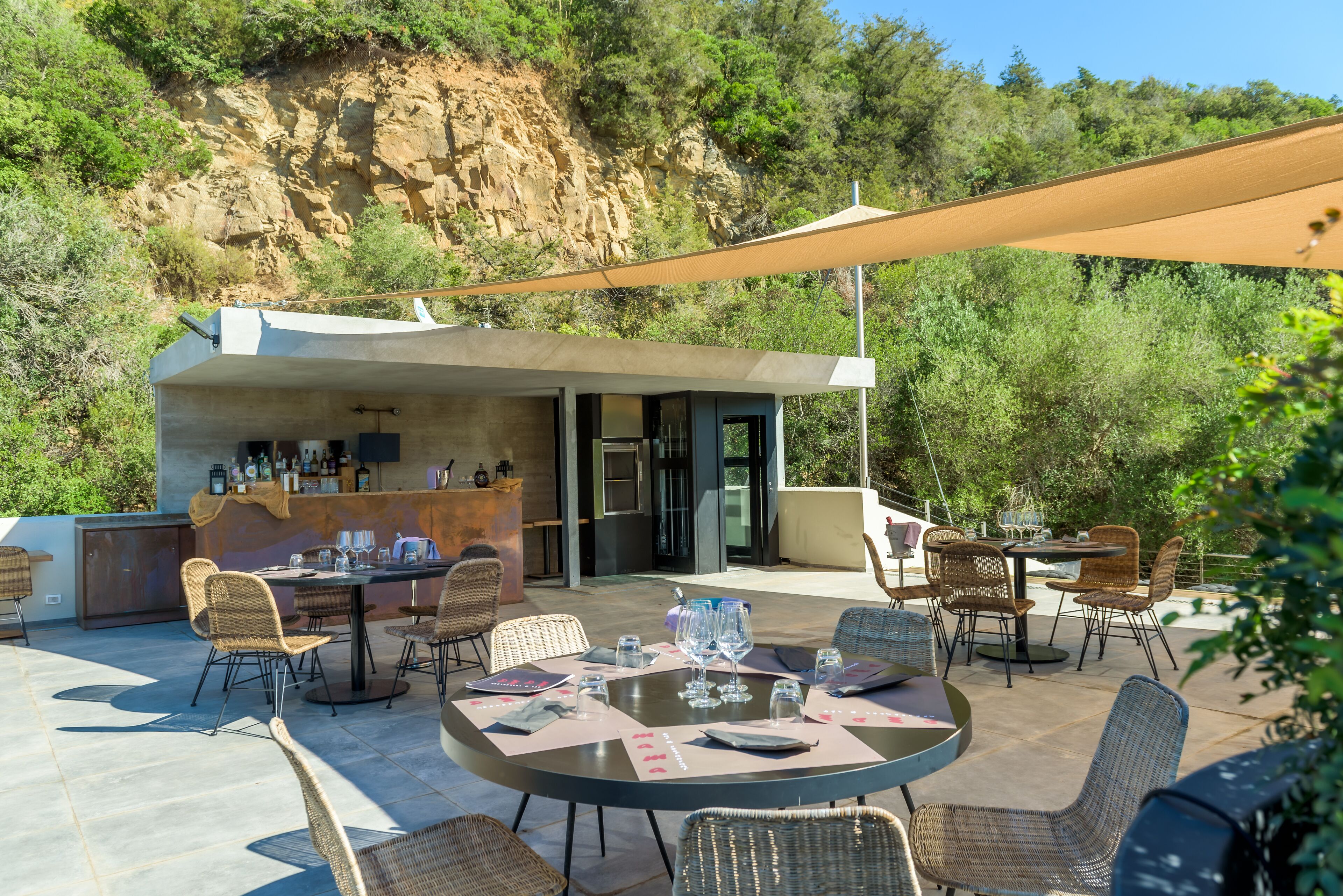 Un patio al aire libre con mesas redondas y sillas de mimbre, una zona de bar y vistas a una ladera rocosa.